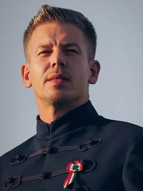 Low-angle golden-hour portrait of Péter Magyar at Heroes’ Square in Budapest on 15 March 2026, wearing a dark braided Hungarian formal jacket and national cockade; the…